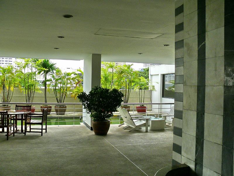 P1000033.JPG - As you emerge from the elevator bank on the fourth floor you pass an interior fountain and emerge into this open space. At the eastern edge (background) is a reflecting pool. Tall potted plants provide a graceful accompaniment to the verticality of highrise condominiums beyond. The shade provided by the condo tower provides a cool place to lounge at picnic tables and recliners.